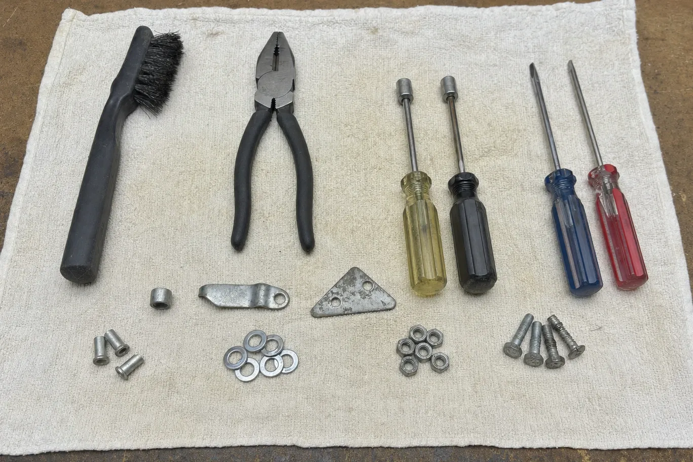 Wire brush, pliers, nut drivers and screwdrivers above sorted washers, nuts and screws on a cloth